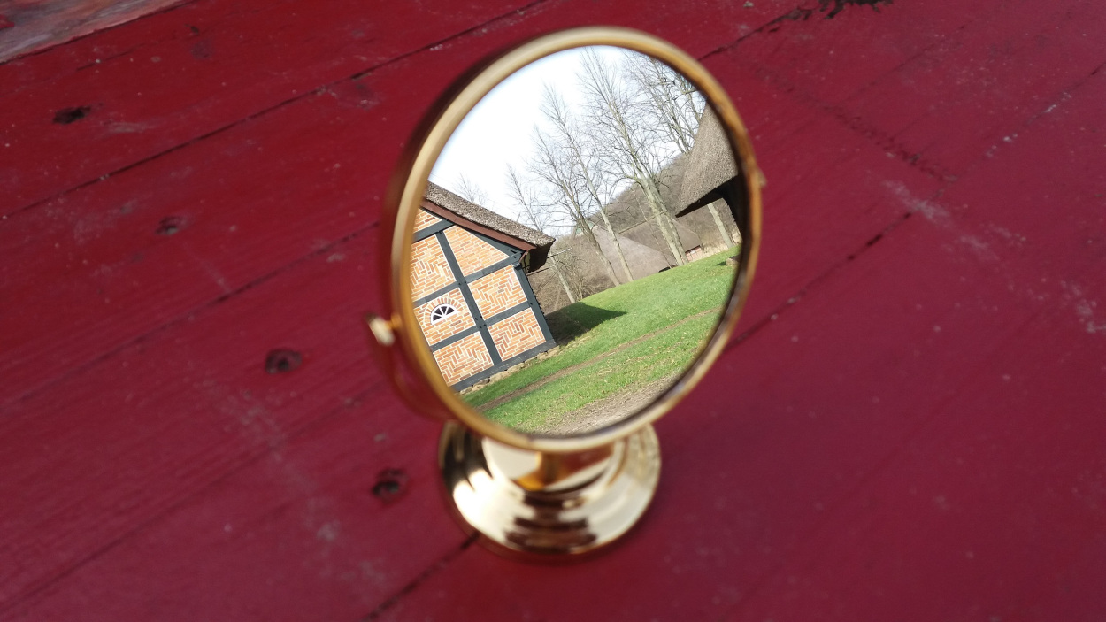 Ein golderner Spiegel auf roter Oberfläche. Im Spiegelbild ist ein Teil von Molfsee Gelände zu sehen.
