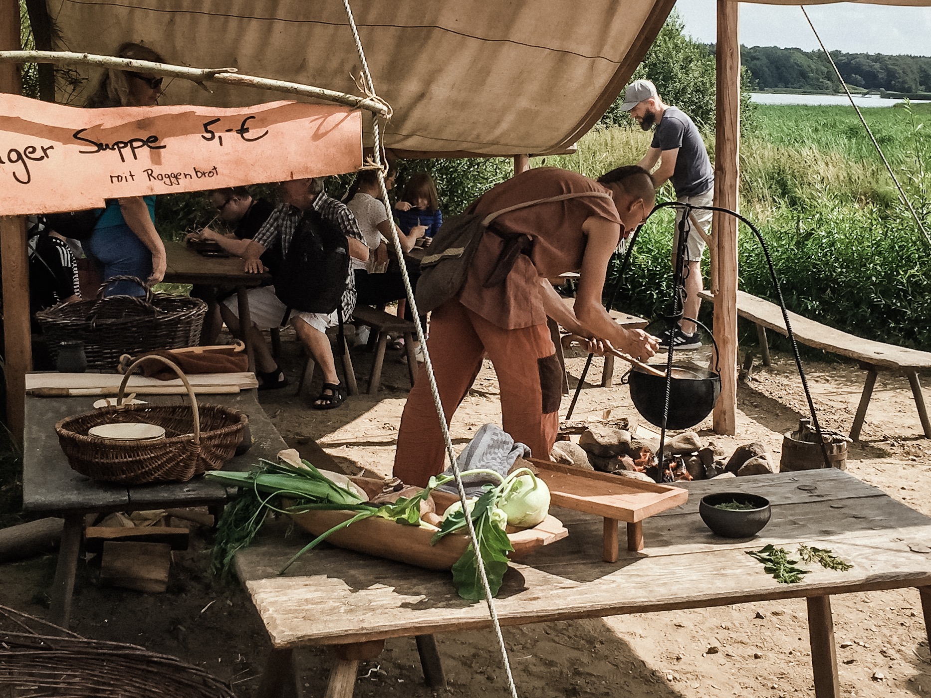 Menschen in einem Zeltlager mit Holztischen und Bänken auf dem Gelände des Wikinger Museums Haithabu.