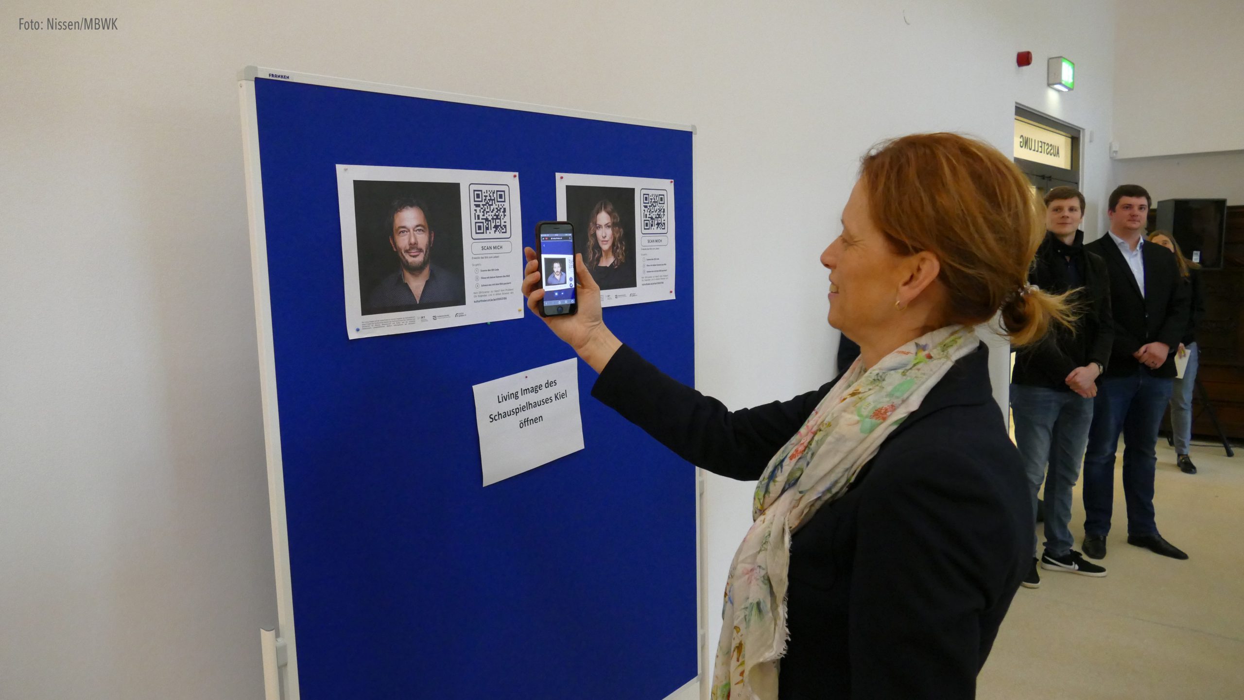 Eine Frau steht vor einer Pinnwand und sieht sich mit ihrem Smartphone Living Images an.